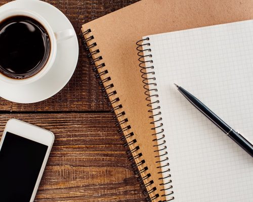 Pen perched on top of two notebooks, next to a cup of coffee and a smartphone